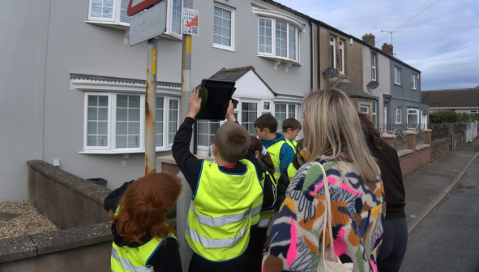Children wearing high-visibility vests gather around an orienteering control marker attached to a street sign on a residential road, while accompanying adults observe. The group is taking part in an outdoor orienteering activity in a village setting with houses lining the street.