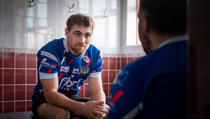 A rugby player sat in a changing room talking to another player who is sat opposite with his back to the camera